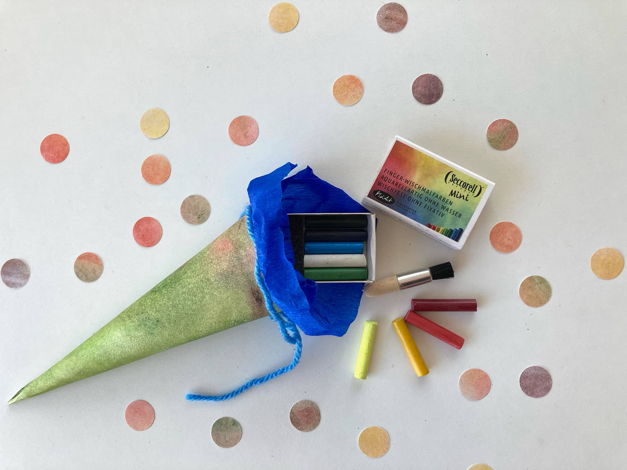 Kleine Schultüte mit Seccorell-Farben und Bürstchen, umgeben von bunten Papierkonfetti.